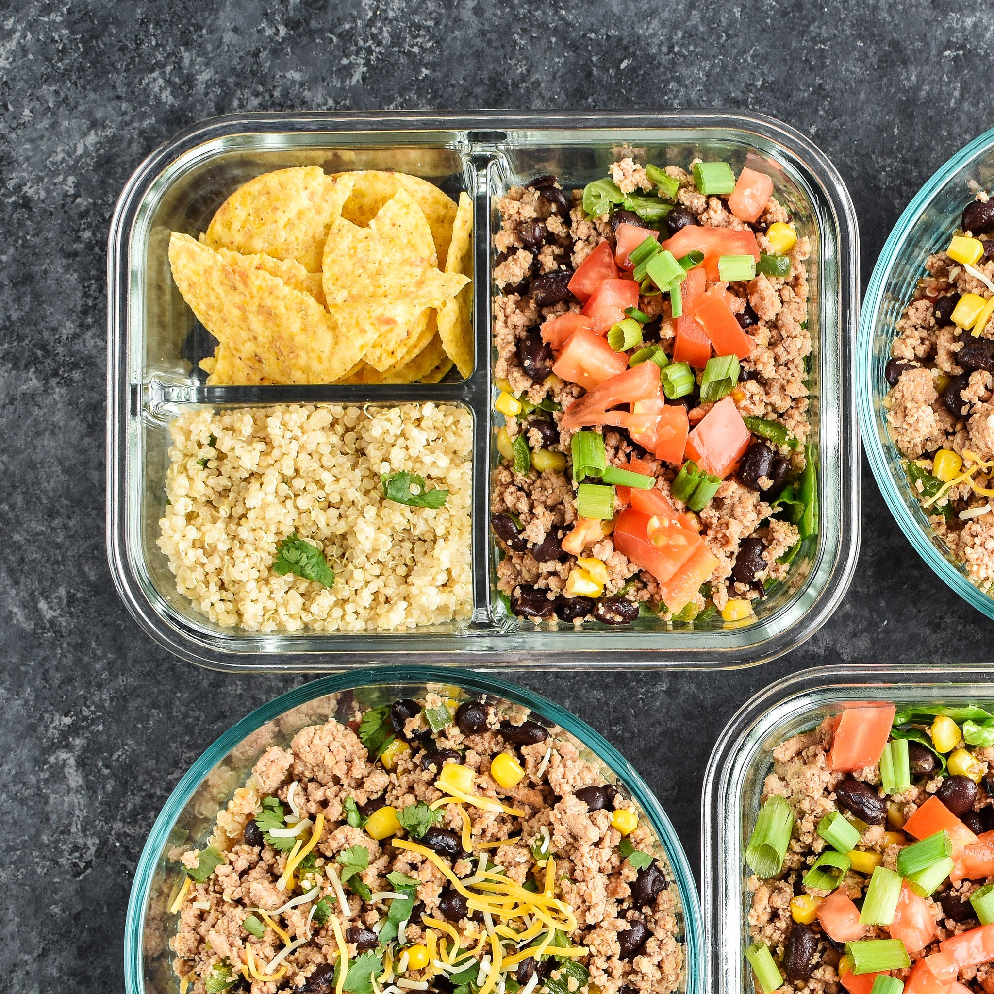 Meal prepped lunches viewed from above for Hot & Cold Turkey Taco Meal Prep; two taco salads with quinoa and chips on the side and two taco bowls with tomato and green chili quinoa.