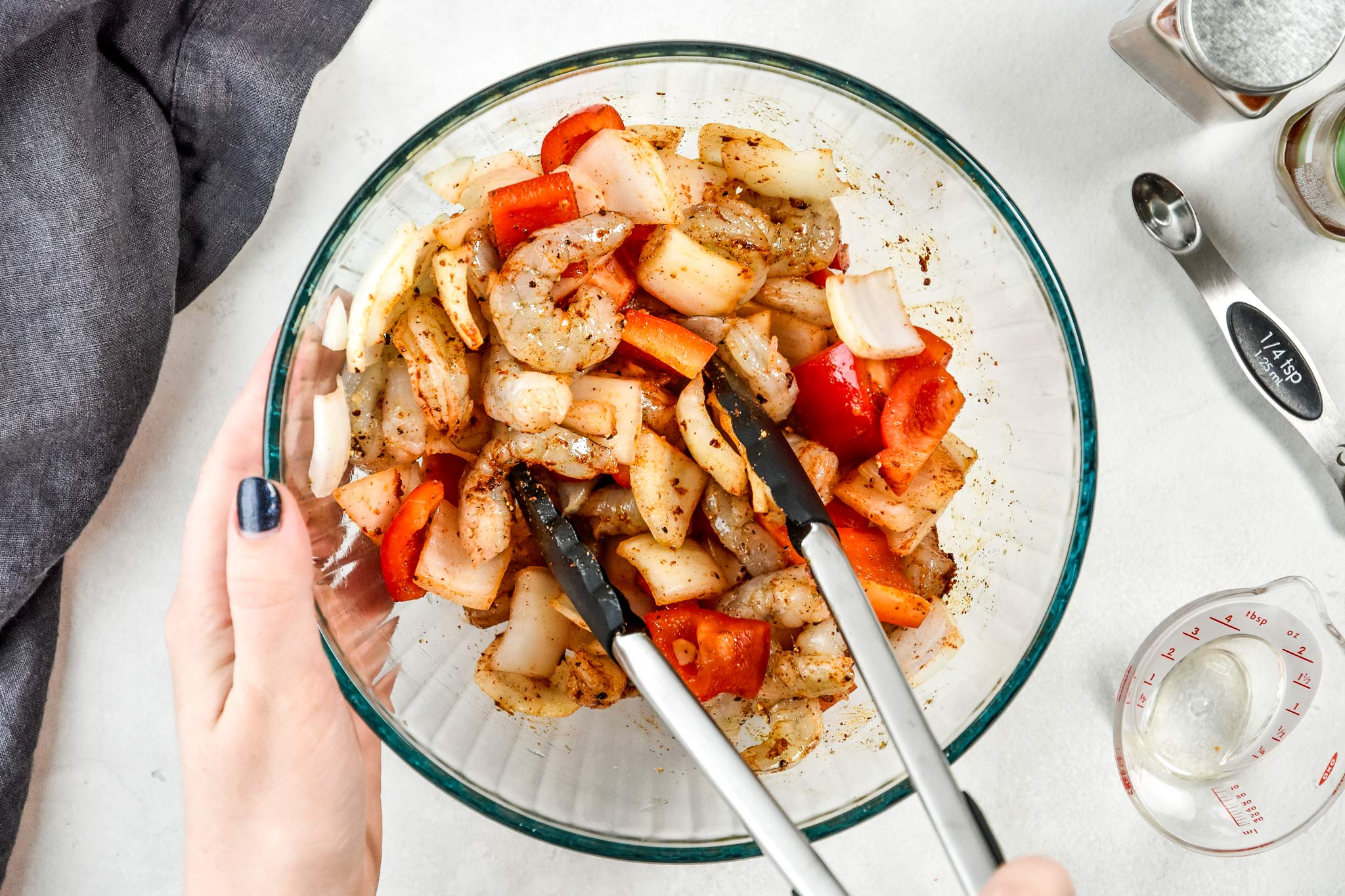 using tongs to stir the spices, shrimp and veggies in a bowl