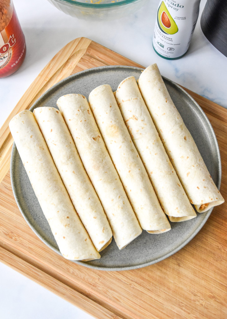 six rolled air fryer buffalo chicken taquitos on a plate before cooking.