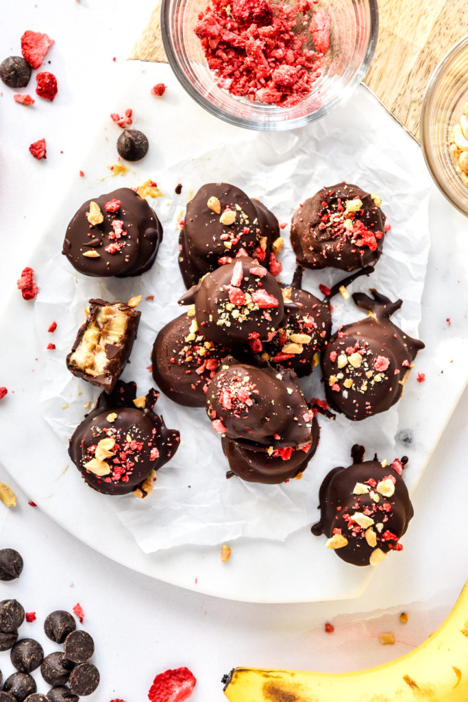 chocolate covered pb&j banana bites with peanuts and freeze dried strawberry crumbs on top on a cutting board.