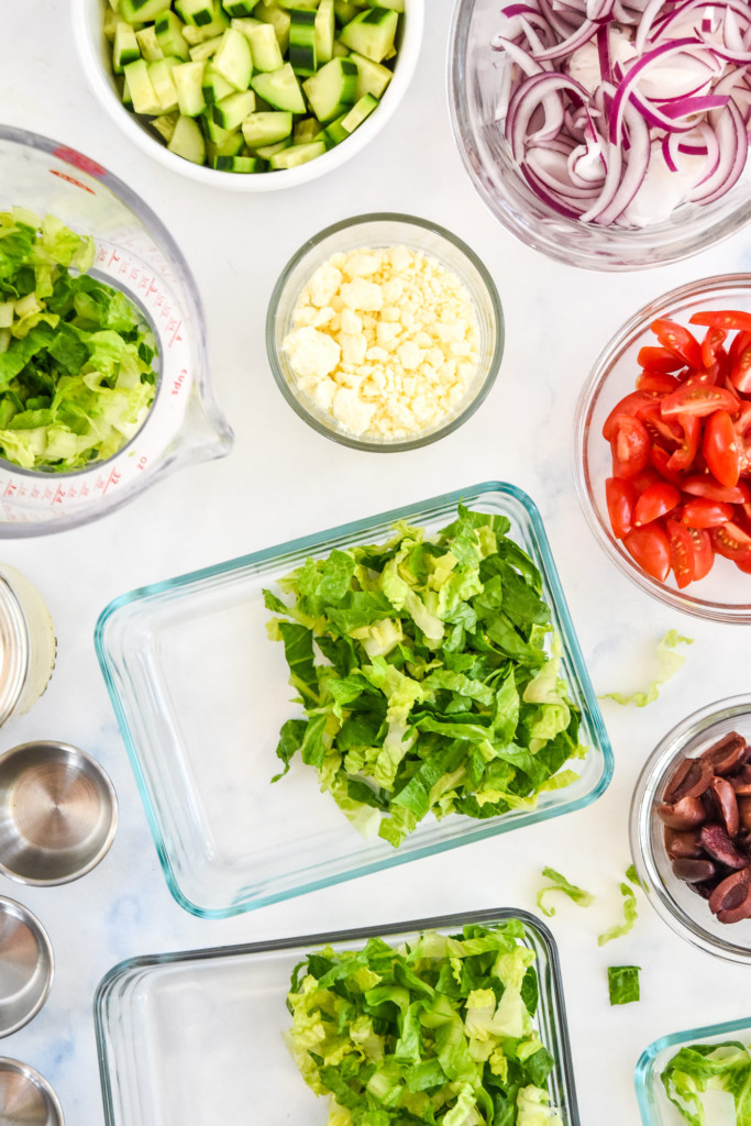 adding romaine and assembling greek chicken salad meal prep in glass containers.