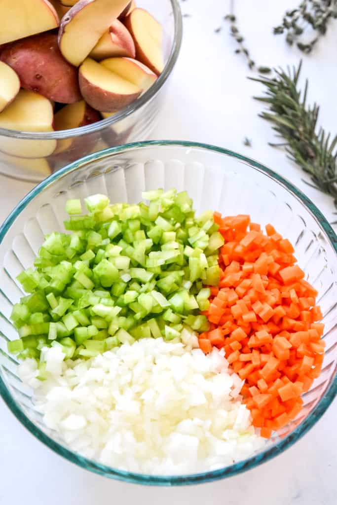 diced onions carrots and celery in a large glass bowl with red potatoes in the background.