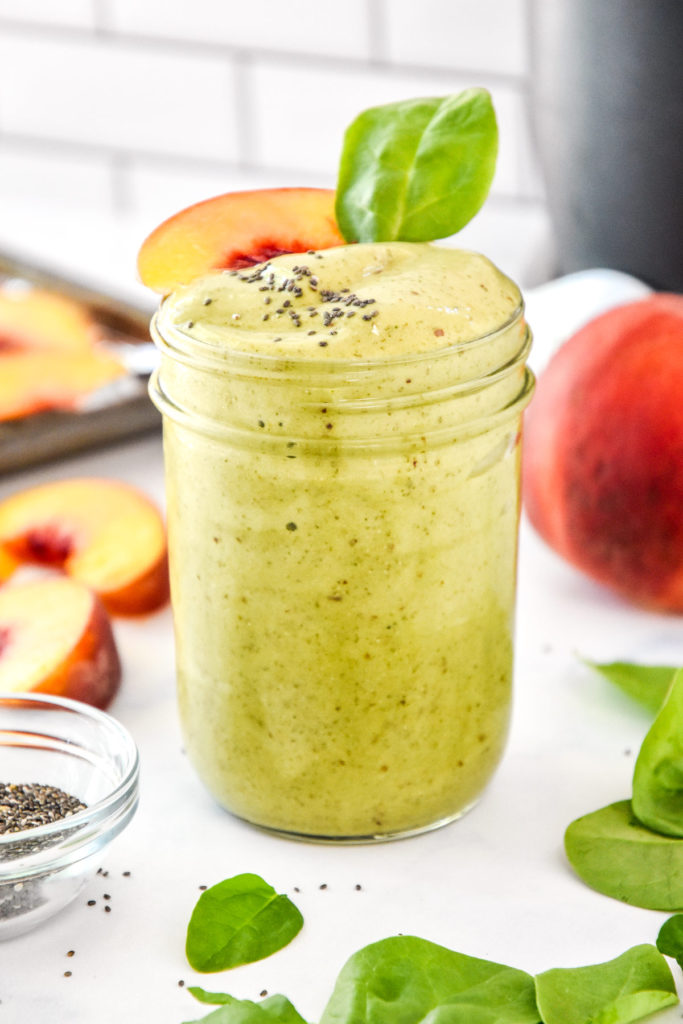 Peaches and green protein smoothie in a glass mason jar with a peach slice on top.