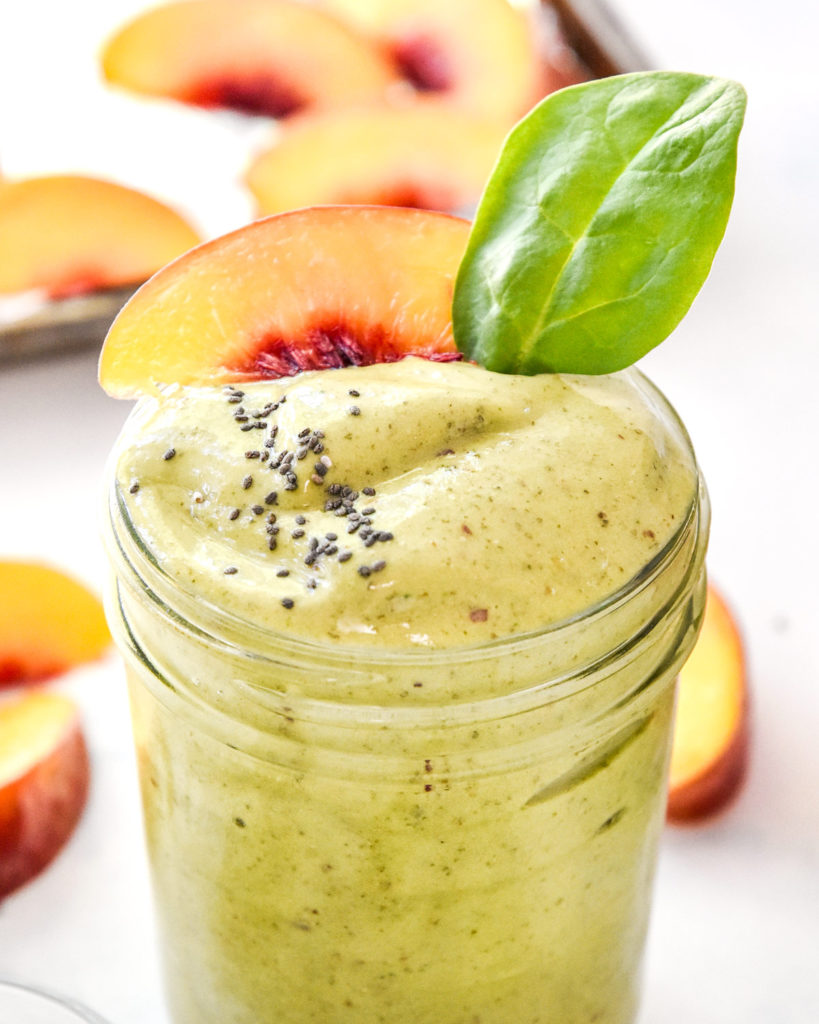peaches and green protein smoothie with spinach leaf, chia seeds and peach slice on top.