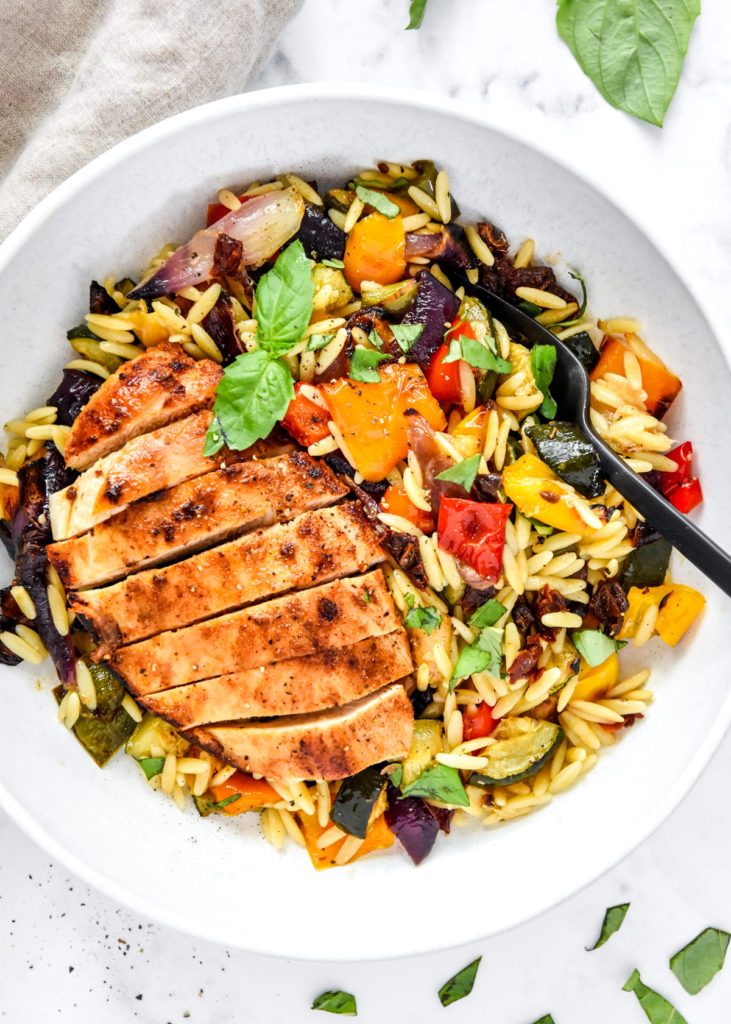 roasted vegetable orzo salad with sliced chicken breast in a white bowl.