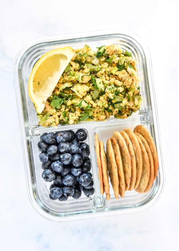 herby lemon mashed chickpea salad in a glass meal prep container with blueberries and crackers.